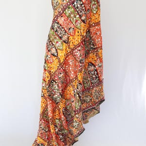 May include: A vibrant, patterned textile in shades of orange, yellow, red, green, and black. The fabric is adorned with intricate floral and geometric designs, and embellished with small, reflective accents. The edges are finished with a dark border.