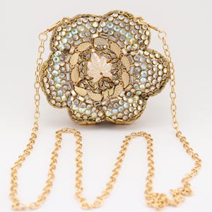 May include: A flower-shaped clutch bag featuring a gold-tone metal frame adorned with iridescent, circular crystals and small gold leaf-shaped accents. The bag includes a long, thin gold-tone metal chain strap attached to the top sides.
