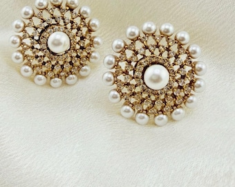 May include: A pair of circular stud earrings featuring a central white pearl surrounded by a gold-toned metal frame encrusted with small clear crystals and an outer border of evenly spaced white pearls arranged in a sunburst pattern.