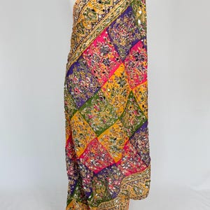 May include: A colorful, patterned scarf with a patchwork design. The scarf features squares of pink, purple, green, and yellow, adorned with intricate embroidery and small, reflective mirror accents. The edges are trimmed with a detailed gold border.