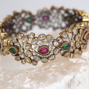 May include: A gold-toned bracelet with intricate floral designs. The bracelet features clear, round gemstones and is accented with oval-shaped red and green gemstones. The jewelry is displayed on a white surface.