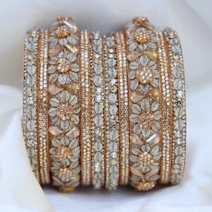 May include: A set of gold-toned bangles featuring intricate floral patterns and leaf motifs. The surface is embellished with numerous small clear crystals and larger peach-colored teardrop stones, creating a textured, metallic appearance.