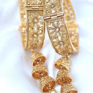 May include: Two gold-colored bangles with floral patterns and small clear stones. Each bangle has a series of bell-shaped charms hanging from it, with a small pearl at the end of each charm. The bangles are on a white background.