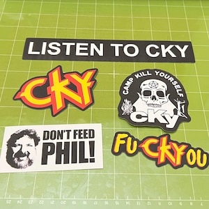 May include: A collection of black and white stickers with text and graphics. One sticker reads "LISTEN TO CKY". Other stickers include "CKY" in yellow and red, a skull with "CAMP KILL YOURSELF", "DON'T FEED PHIL!", and "FUCK YOU".