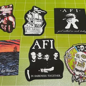 May include: A collection of AFI band stickers. The stickers feature various designs, including a fist with the text "ALL FOR YOU", a ship, band members, and a skull with a pirate hat. Some stickers have text like "Black Sails in the Sunset" and "IN DARKNESS TOGETHER..."