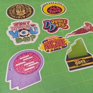 May include: A collection of colorful stickers featuring various designs and text. Designs include a Mayan-style face, the words "ALL THAT" in yellow, "WHAT WOULD YOU DO?", "DOUBLE DARE", a reclining figure, "NICKELODEON ARCADE", a brain, and a "GUTS" logo.