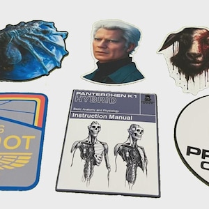May include: A collection of stickers featuring various designs. Included are a blue, textured creature, a portrait of a man, a goat with blood-like streaks, a patch with text "USCSS MAGINOT", a manual with anatomical illustrations, and a Prodigy Corp sticker.