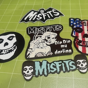 May include: A collection of Misfits band stickers in various shapes. Designs include the band's logo in a bat shape, a skull, and a skull with an American flag design. One sticker reads "Die Die mu darling".