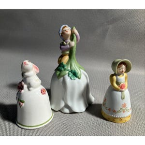 May include: Three ceramic figurines: a white bunny, a boy climbing a flower, and a girl holding flowers. The bunny figurine has pink accents. The boy is dressed in purple and yellow. The girl wears a yellow dress and a bonnet. All are bell-shaped.