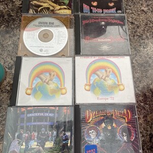 May include: A collection of Grateful Dead CDs, featuring album art with various designs. Titles include "In the Dark," "Europe '72," and "Dozin' at the Knick." Some cases have handwritten notes. The CDs are in their original jewel cases.