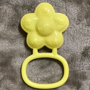 May include: A yellow plastic baby rattle shaped like a flower with a circular handle. The flower has five petals, each with a small flower design. The center of the flower has the letters "CPK".