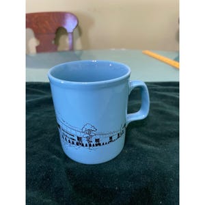 May include: A light blue ceramic mug with a black and white graphic of a building or stadium. The mug has a handle and a simple, clean design. The mug is likely for beverages.