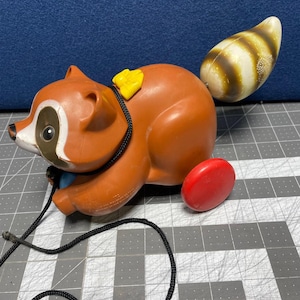 May include: A brown and black pull toy raccoon with a yellow tag on its back. The raccoon has a white stripe on its face and a striped tail. The toy has red wheels and a black pull string.