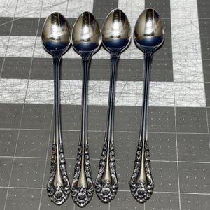 May include: Four silver spoons with long, slender handles and ornate floral designs on the spoon bowls. The spoons are arranged vertically, reflecting light and creating a shiny appearance. The spoons are likely for serving coffee or tea.