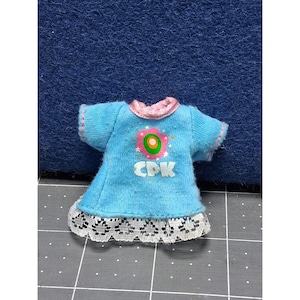 May include: A light blue doll dress with short sleeves and a white lace hem. The dress has a pink collar and sleeve trim, and a flower graphic with the letters "CDK" on the front.