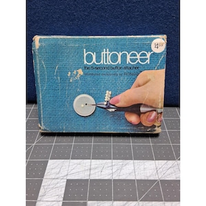 May include: Vintage "Buttoneer" box, a 5-second button attacher. The box is a textured teal color with the product name in white. The image on the box shows a hand using the tool to attach a button. The box is distributed exclusively by RONCO.