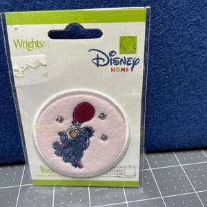 May include: A Disney Home patch featuring Eeyore holding a red balloon, surrounded by small yellow bees, on a pink background. The patch is round with a white border and is packaged in a clear plastic sleeve with the Wrights and Disney Home logos.