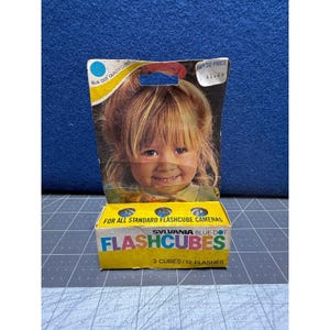 May include: Vintage Sylvania Flashcubes box. The yellow box features the text "FLASHCUBES" in blue and green, with a picture of a smiling child. The box is labeled "3 CUBES/12 FLASHES" and "FOR ALL STANDARD FLASHCUBE CAMERAS".