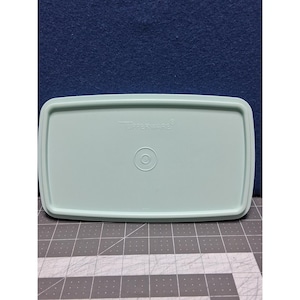 May include: A light green Tupperware rectangular lid. The lid has rounded corners and a slightly raised edge. The Tupperware logo is visible on the top surface, along with a circular indentation in the center.