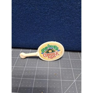 May include: A vintage Cabbage Patch Kids Cornsilk Kids toy mirror. The mirror is oval-shaped with a white handle. The mirror's face features the Cabbage Patch Kids logo and the words "Cornsilk Kids" in a circular design.