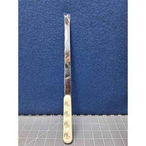 May include: A long, thin, silver-colored metal knife with a cream-colored handle featuring a repeating pattern of small teddy bears. The blade is encased in a clear plastic sleeve, and the word COLLECTION is printed vertically on the metal blade.