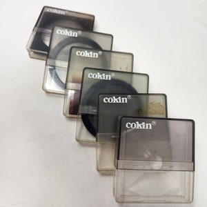 May include: Five clear plastic cases with black circular filters inside. Each case has the word "cokin" printed on it in white.