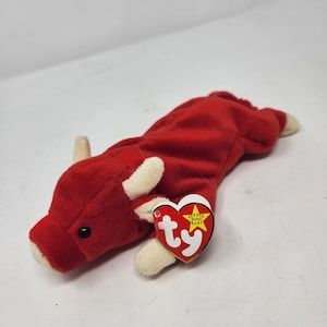 May include: Red plush toy bull with a white face and horns. The toy has a Ty tag attached to it that says "Beanie Original Baby".
