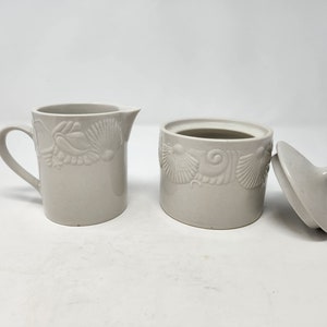 May include: A white ceramic creamer and sugar bowl set with a seashell design. The creamer has a handle and a spout. The sugar bowl has a lid.