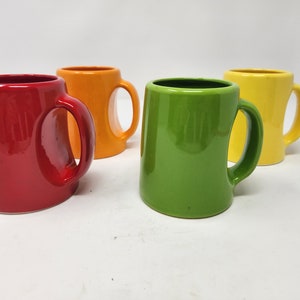 May include: Four ceramic mugs in different colors: red, orange, green, and yellow. Each mug has a unique handle design.