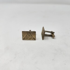 May include: A pair of gold-toned cufflinks with a geometric design. The cufflinks are square-shaped with a triangular pattern in the center.