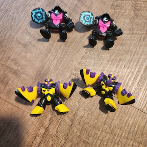 May include: Four small plastic action figures. Two are black with pink accents and blue and white spinning discs. Two are yellow with black and purple accents and wings.