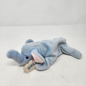 May include: A light blue plush elephant toy with a pink ear and a red heart-shaped tag. The tag has the words "Ty Inc." and "Beanie Babies" printed on it.