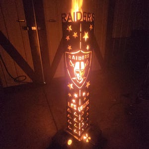 May include: A metal fire pit with a cutout design of the Las Vegas Raiders logo and the words "RAIDERS" and "LAS VEGAS". The fire pit is lit and glowing orange.