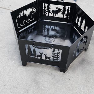 May include: Black metal fire pit with cutouts of a bear, moose, deer, and trees. The fire pit has a handle on the side and sits on four legs.