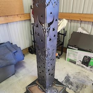 May include: A metal fire pit with a mountain and star design. The fire pit is made of steel and has a square base. The fire pit is decorated with a horseshoe, a bull skull, and stars. The fire pit is made by DB Designs.
