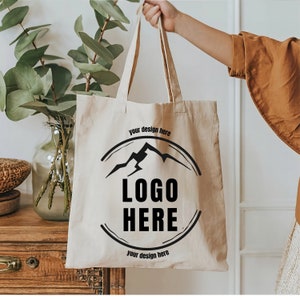 May include: A beige tote bag with black handles and a black logo design featuring a mountain range and the text "LOGO HERE" with the words "your design here" above and below the logo.