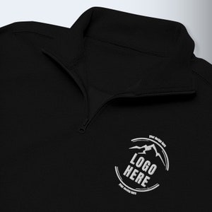 May include: Black quarter-zip fleece pullover with a white embroidered logo that says "LOGO HERE" and has a mountain design. The text "your design here" appears above and below the logo.