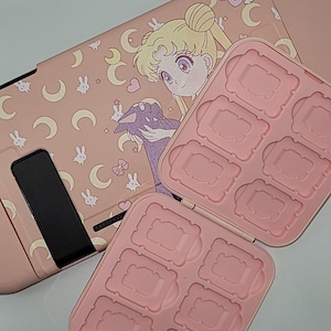 Può includere: Custodia rosa per Nintendo Switch con un design di Sailor Moon. La custodia ha un portacarte di gioco integrato con otto slot.