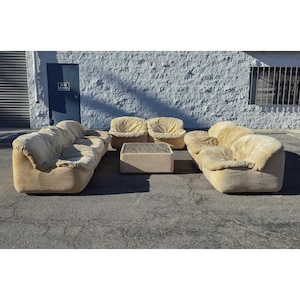 May include: A beige upholstered living room set, including a sofa, two armchairs, and a square coffee table. The furniture has a soft, rounded design. The set is placed outdoors against a white wall.