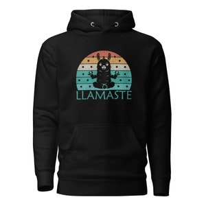 Op de afbeelding: Zwarte hoodie met een vintage-stijl afbeelding van een lama in een yogapose. De afbeelding is geplaatst tegen een zonsondergang achtergrond met sterren en de tekst "LLAMASTE".