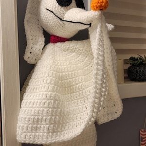 Puede incluir: Un Zero de ganchillo hecho a mano de Pesadilla antes de Navidad. El perro fantasma blanco tiene ojos negros, una nariz larga y un collar rojo. Sostiene una calabaza naranja. La figura de ganchillo está colgada en una pared.