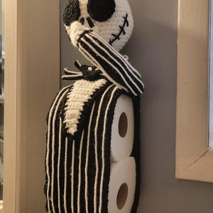 May include: Crocheted toilet paper holder in the shape of Jack Skellington from The Nightmare Before Christmas. The holder is white with black accents, including a striped suit and facial features. It holds two rolls of toilet paper.