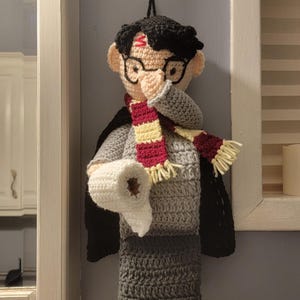 May include: Crocheted Harry Potter-themed toilet paper holder. The holder features a black-haired character with glasses, a red and gold scarf, and a lightning bolt scar. The holder is gray and black, holding a roll of white toilet paper.