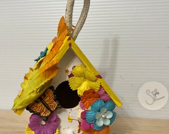 May include: A small, white birdhouse with a yellow roof and a rope hanger. The birdhouse is decorated with colorful paper flowers, a butterfly, and white paint splatters.