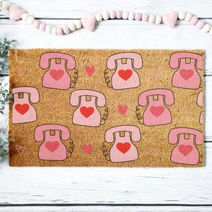 May include: A brown coir doormat with a pink and red pattern of retro rotary phones with hearts. The doormat has a beige background with a subtle texture.