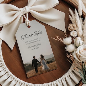 Puede incluir: Una etiqueta de recuerdo de boda crema con las palabras "Thank You" en escritura elegante. La etiqueta presenta una foto de una pareja tomados de la mano, con un fondo de montaña. Está atada con un lazo color crema y descansa sobre una bandeja de madera con detalles florales.