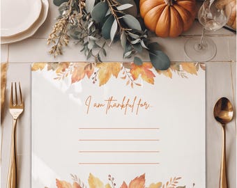 Könnte beinhalten: Ein weißer Tischset mit einem Herbstblatt-Design und dem Text "I am thankful for...". Das Tischset ist von goldenem Besteck, einem Kürbis und Herbstlaub umgeben, was eine festliche Thanksgiving-Tischdekoration schafft.