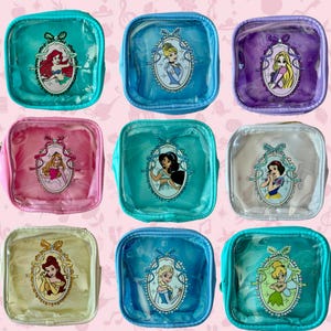 May include: Nine clear, square cosmetic bags with different Disney princess portraits. Each bag has a colored border and a zipper. The princesses include Ariel, Cinderella, Rapunzel, Aurora, Jasmine, Snow White, Belle, Elsa, and Tinkerbell.