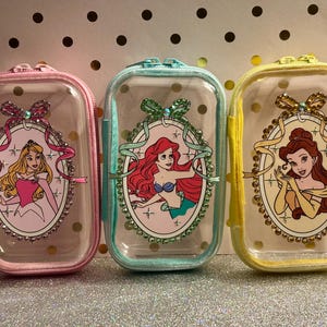 May include: Three clear, rectangular zippered pouches with princess illustrations. The pouches are pink, teal, and yellow, each featuring a different princess in an oval frame with decorative embellishments. The pouches have a clear front and a solid color border.