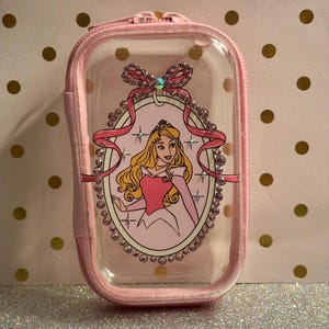 May include: Pink and clear zippered pouch with a princess illustration. The pouch has a pink zip and trim, with a clear front panel displaying a princess in a pink dress. Embellished with rhinestones and a bow.